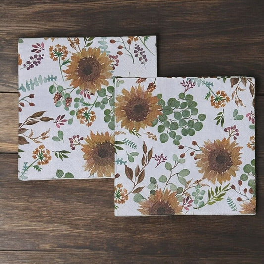 Cocktail-size paper napkin featuring a watercolor botanical print of golden sunflowers, eucalyptus sprigs, dusty rose berries, and wildflower stems on a soft white background, photographed on a warm wood surface.
