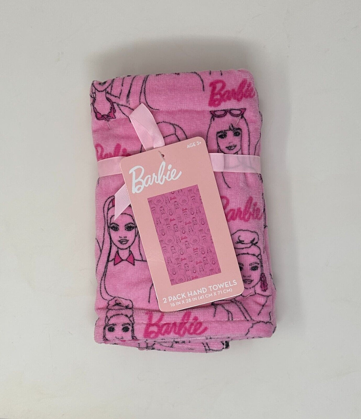 Two pink hand towels with an all-over pattern featuring Barbie and friends, rectangular in shape, laid flat with a product tag attached.
