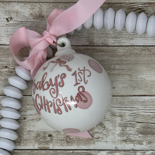 White ornament with 'Baby's 1st Christmas' text and pink ribbon on a wooden surface.