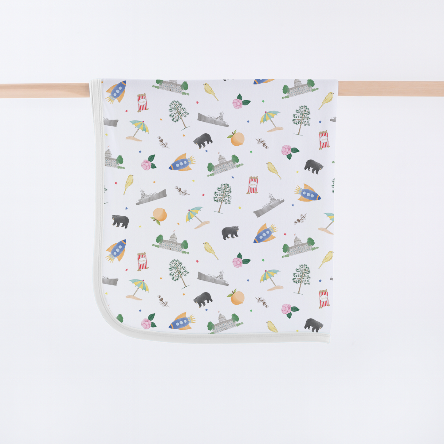 A square white baby blanket with rounded corners, featuring a colorful cartoon pattern of rockets, trees, owls, bears, and houses, hanging from a light wood dowel against a white background.