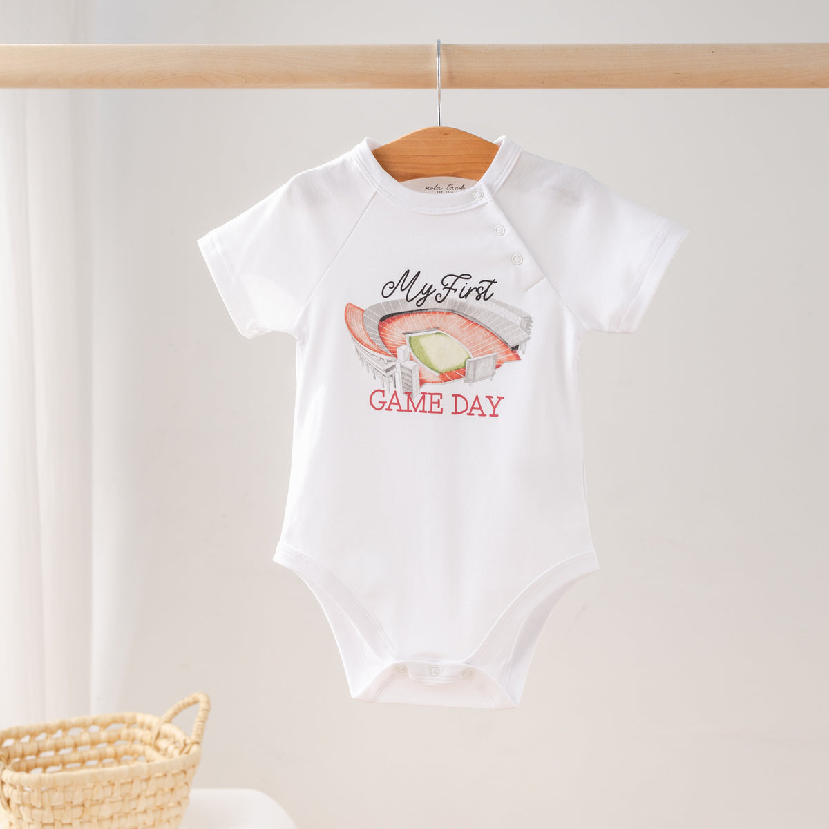 White baby onesie with 'My First Game Day' text and graphics on a wooden hanger.
