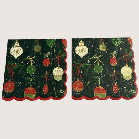 Cocktail-size decoupage paper napkin featuring an all-over print of hanging Christmas ornaments in deep red forest green cream and gold with gold foil detailing suspended from pine sprigs with ribbon bows on a deep forest green background with gold star accents and a red scalloped edge border, photographed on a light grey surface.