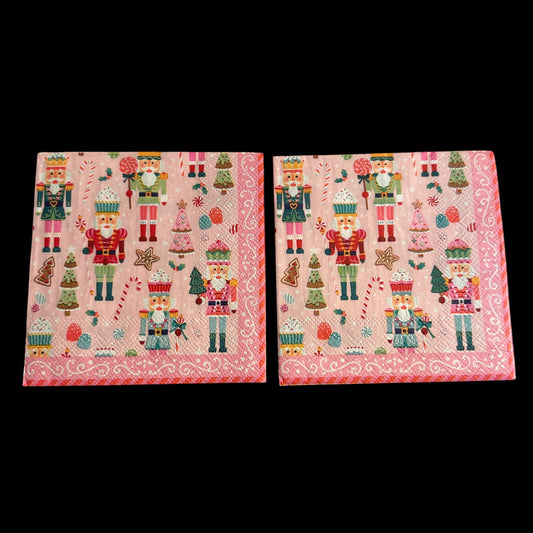 Cocktail-size decoupage paper napkin featuring a lively all-over print of colorful nutcracker soldiers in teal red green and pink uniforms with candy canes lollipops cupcake nutcrackers gingerbread trees pink Christmas trees and star cookies on a soft bubblegum pink background with a white scrollwork lace border, photographed on a light grey surface.