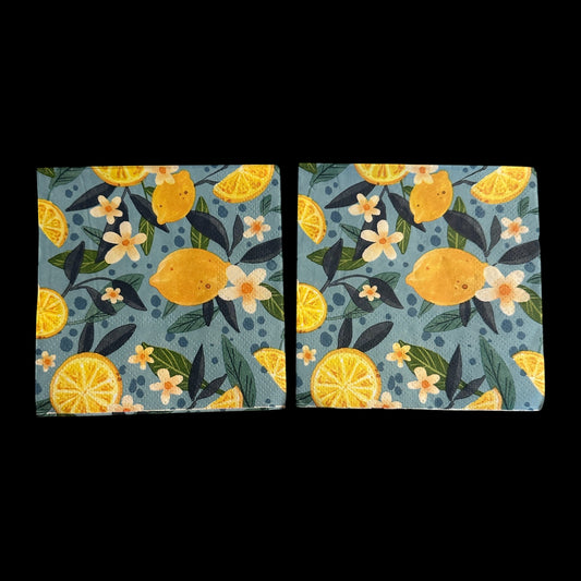 Cocktail-size decoupage paper napkin featuring a lush all-over botanical print of golden whole lemons and halved citrus slices with white orange blossom flowers and rich dark and forest green leaves on a soft dusty blue background with subtle dot accents, photographed on a black surface.