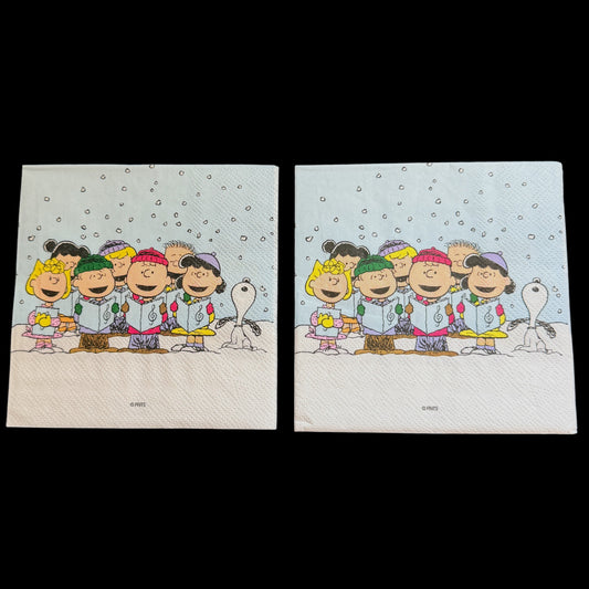 Cocktail size decoupage napkin featuring the Peanuts gang including Charlie Brown Snoopy Lucy Sally and friends caroling together in falling snow on a soft blue winter background set of 2