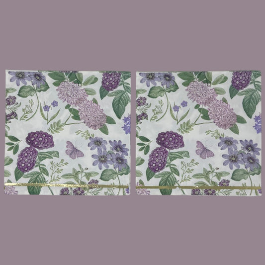Luncheon-size decoupage paper napkin featuring a vintage botanical illustration print of lavender and purple lilac clusters, periwinkle blue anemones, mauve wildflowers, tiny purple butterflies, and green leaves on a white background with gold foil border stripes, photographed against a white wall.