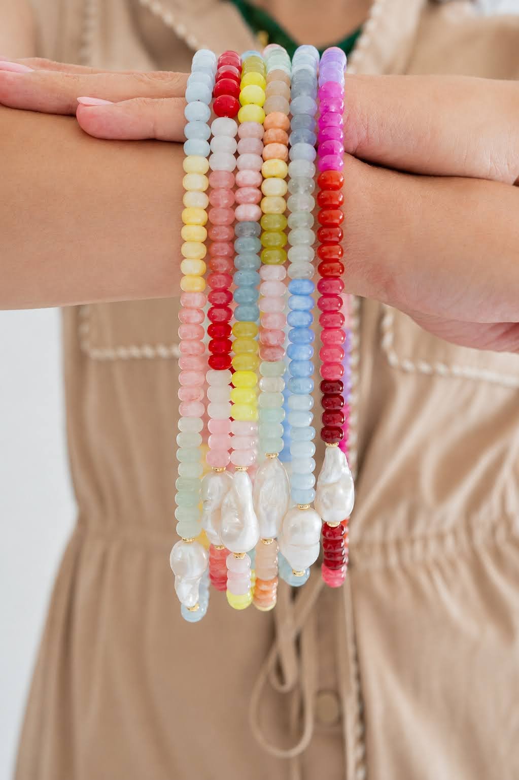 A close-up of a person's hands holding up six colorful beaded bracelets with white baroque pearls, shown against a soft beige background.