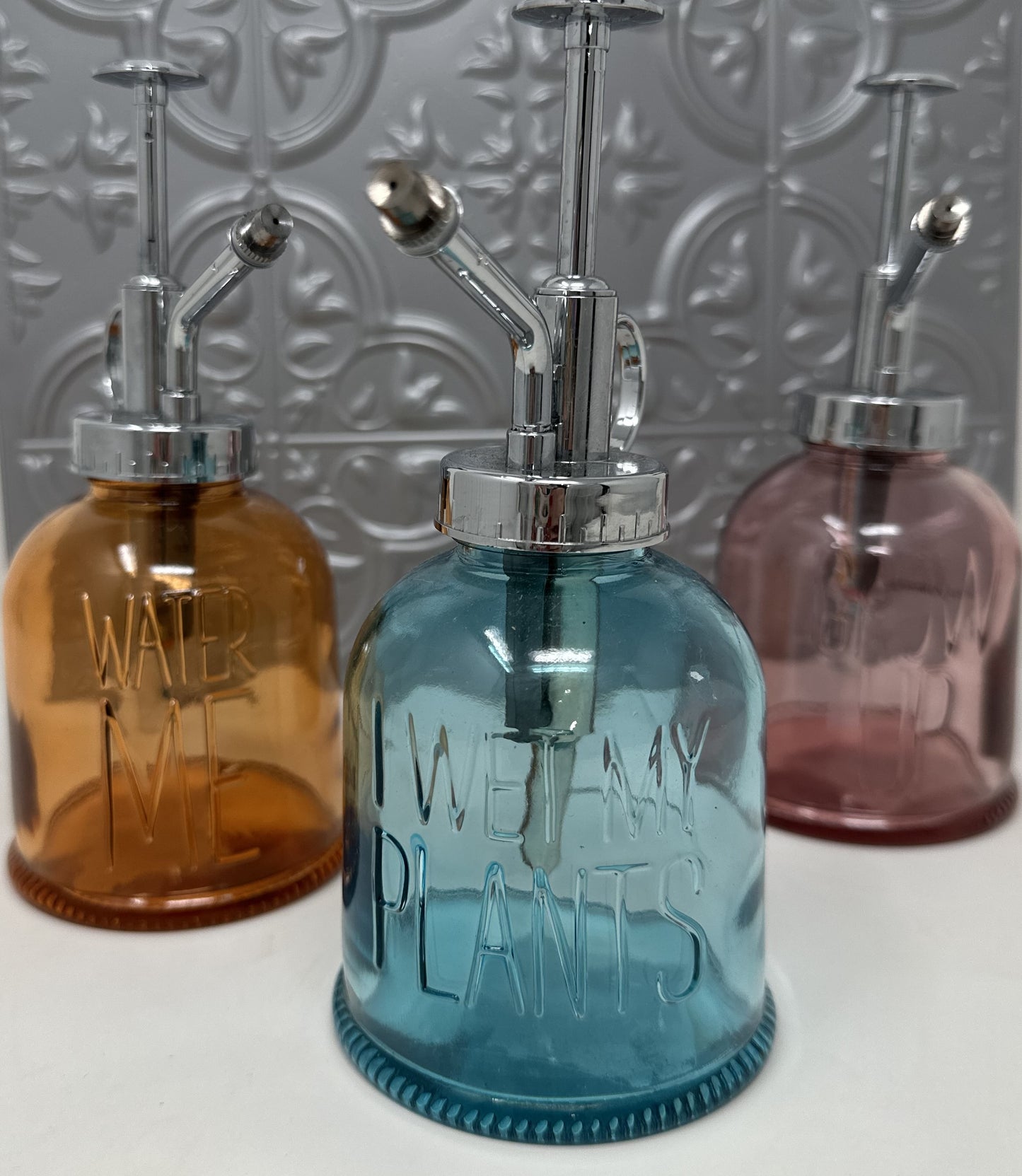 Three Mud Pie tinted glass plant misters in amber, blue, and pink with embossed sayings Water Me, I Wet My Plants, and Grow Up, with chrome pump sprayers, 6.25 x 3.5 inches