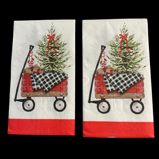 Close-up of two guest-size Christmas napkin featuring a red wagon carrying a decorated Christmas tree, wrapped gifts, and a black-and-white buffalo check blanket on a white background with a red border.