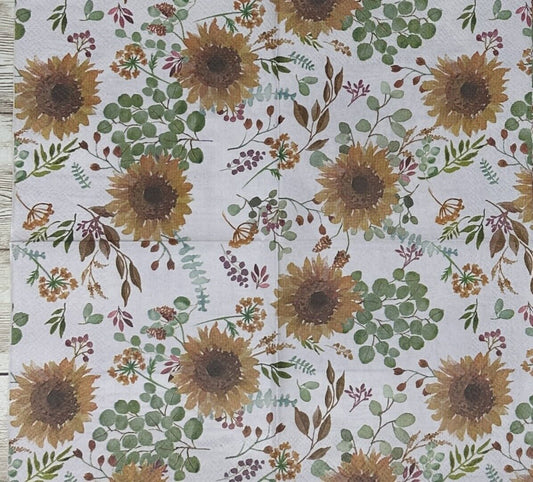 Cocktail-size paper napkin featuring a watercolor botanical print of golden sunflowers, eucalyptus sprigs, dusty rose berries, and wildflower stems on a soft white background, photographed on a warm wood surface.