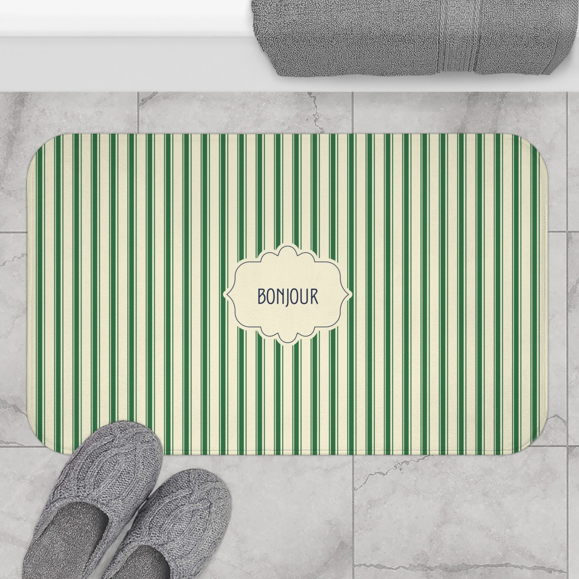 A rectangular bath mat with rounded corners, featuring vertical green and cream stripes and a central cream label that reads 'BONJOUR'. The mat is styled in a bathroom setting on a light gray marble tile floor, with gray slippers at the bottom left, a folded gray towel at the top, and the mat itself centered.