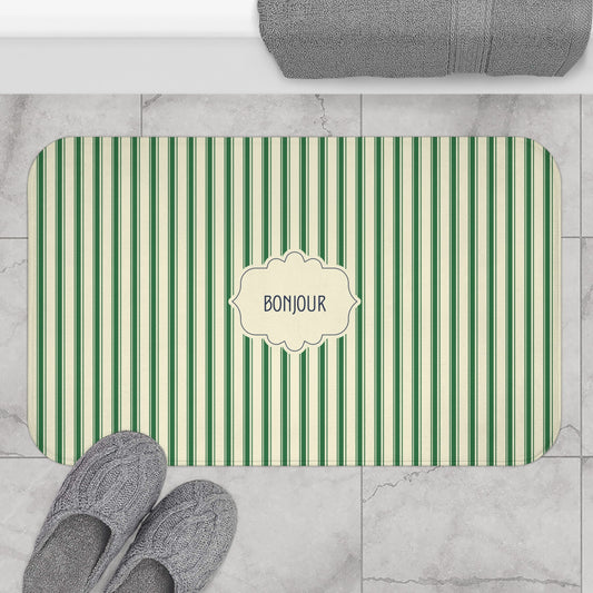 A rectangular bath mat with rounded corners, featuring vertical green and cream stripes and a central cream label that reads 'BONJOUR'. The mat is styled in a bathroom setting on a light gray marble tile floor, with gray slippers at the bottom left, a folded gray towel at the top, and the mat itself centered.