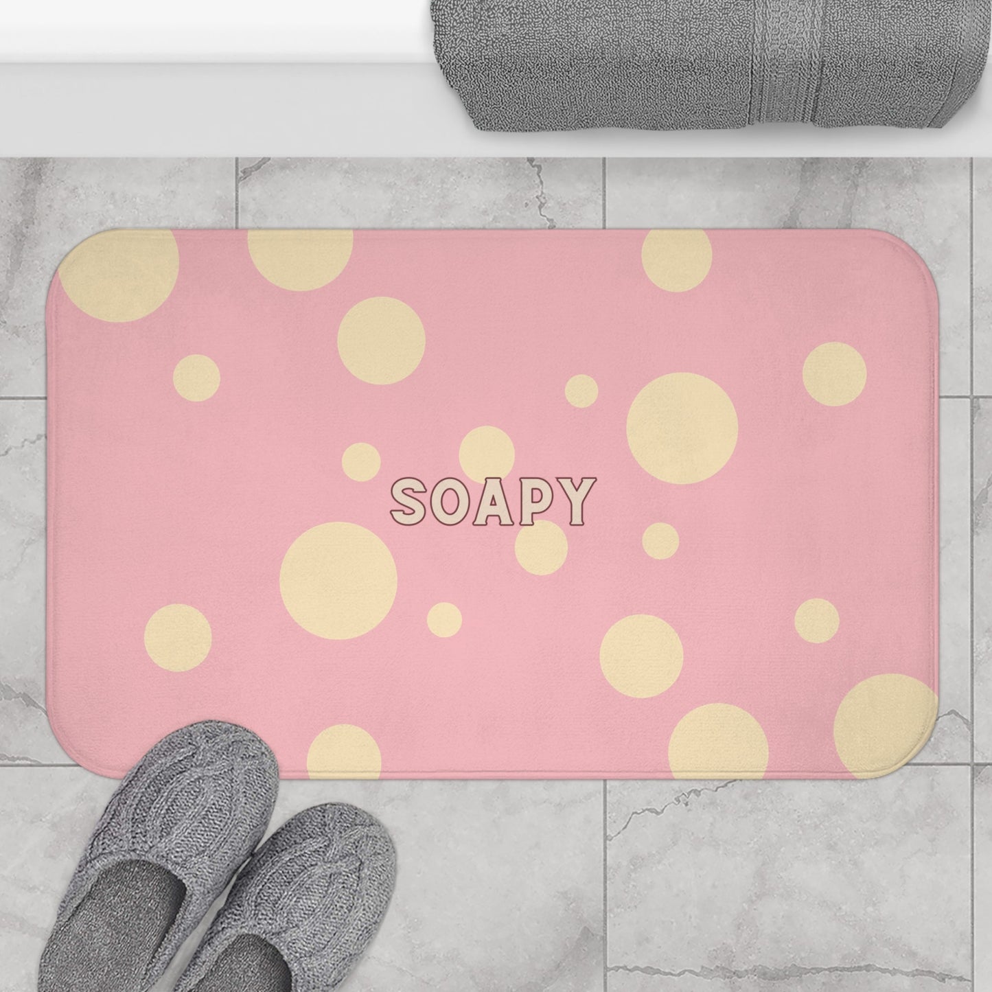 A pink bath mat with a pattern of large and small yellow polka dots and the word 'SOAPY' in the center, shown on a gray marble bathroom floor with gray slippers and a towel.
