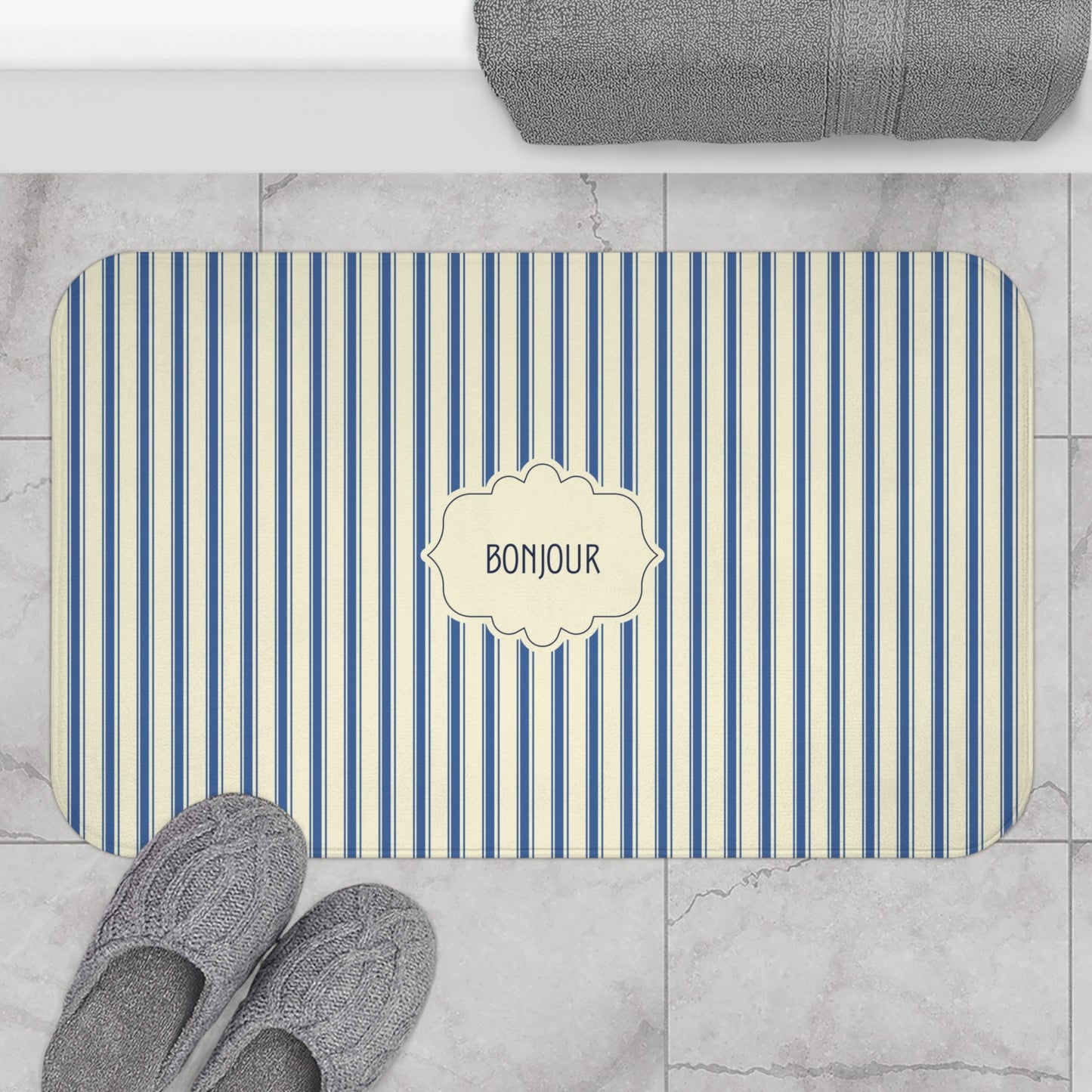 A rectangular bathroom mat with rounded corners, featuring vertical blue and cream stripes and a central cream label that reads 'BONJOUR'. The mat is shown on a light gray marble tile floor next to gray slippers, with a folded gray towel at the top edge.