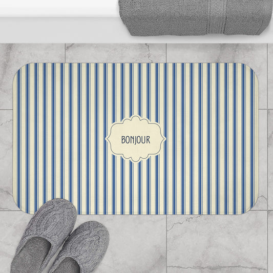 A rectangular bathroom mat with rounded corners, featuring vertical blue and cream stripes and a central cream label that reads 'BONJOUR'. The mat is shown on a light gray marble tile floor next to gray slippers, with a folded gray towel at the top edge.