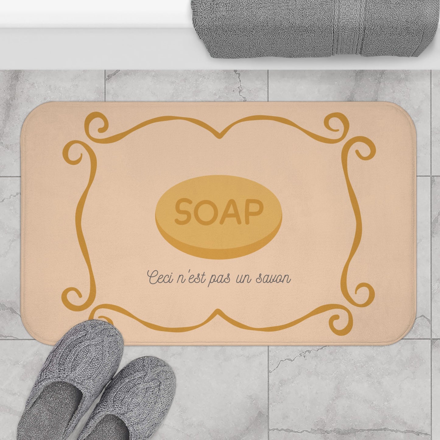 Beige bath mat with gold 'SOAP' design on a marble floor with gray slippers and towel.