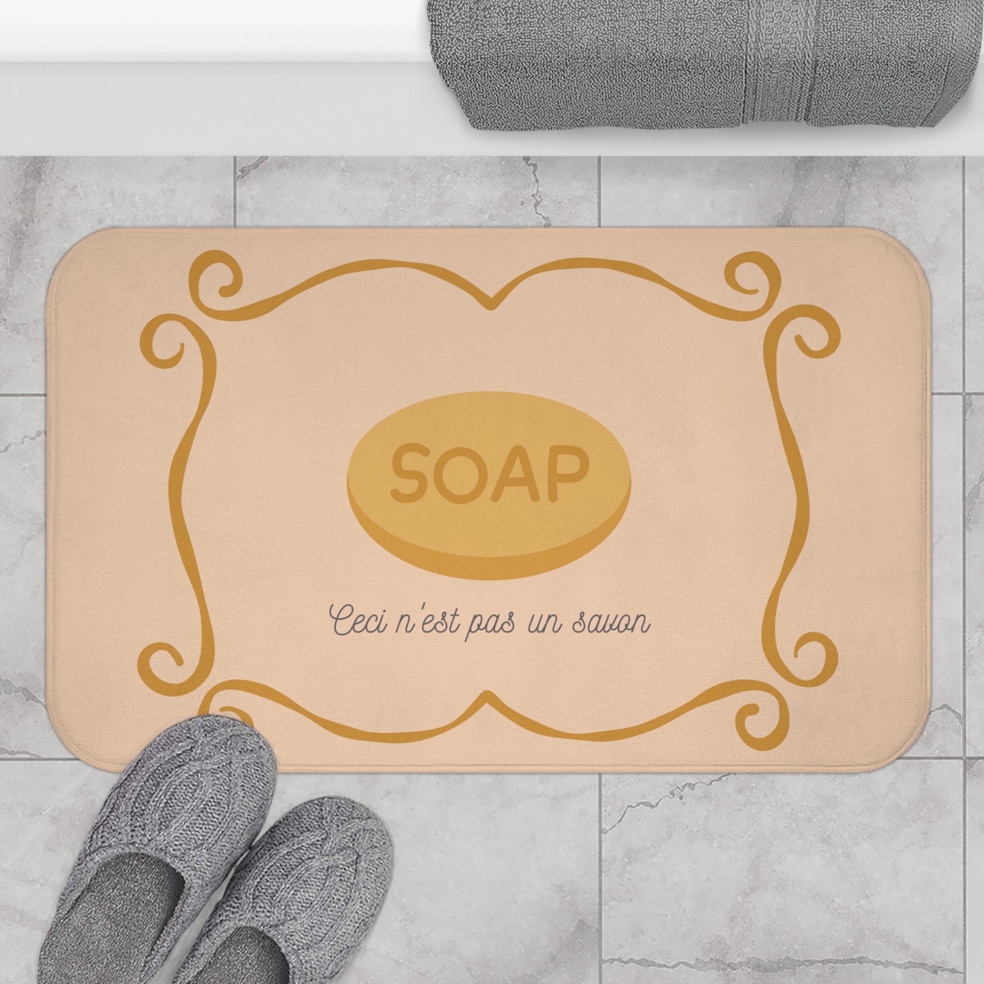 Beige bath mat with gold 'SOAP' design on a marble floor with gray slippers and towel.
