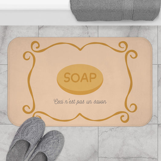 Beige bath mat with gold 'SOAP' design on a marble floor with gray slippers and towel.