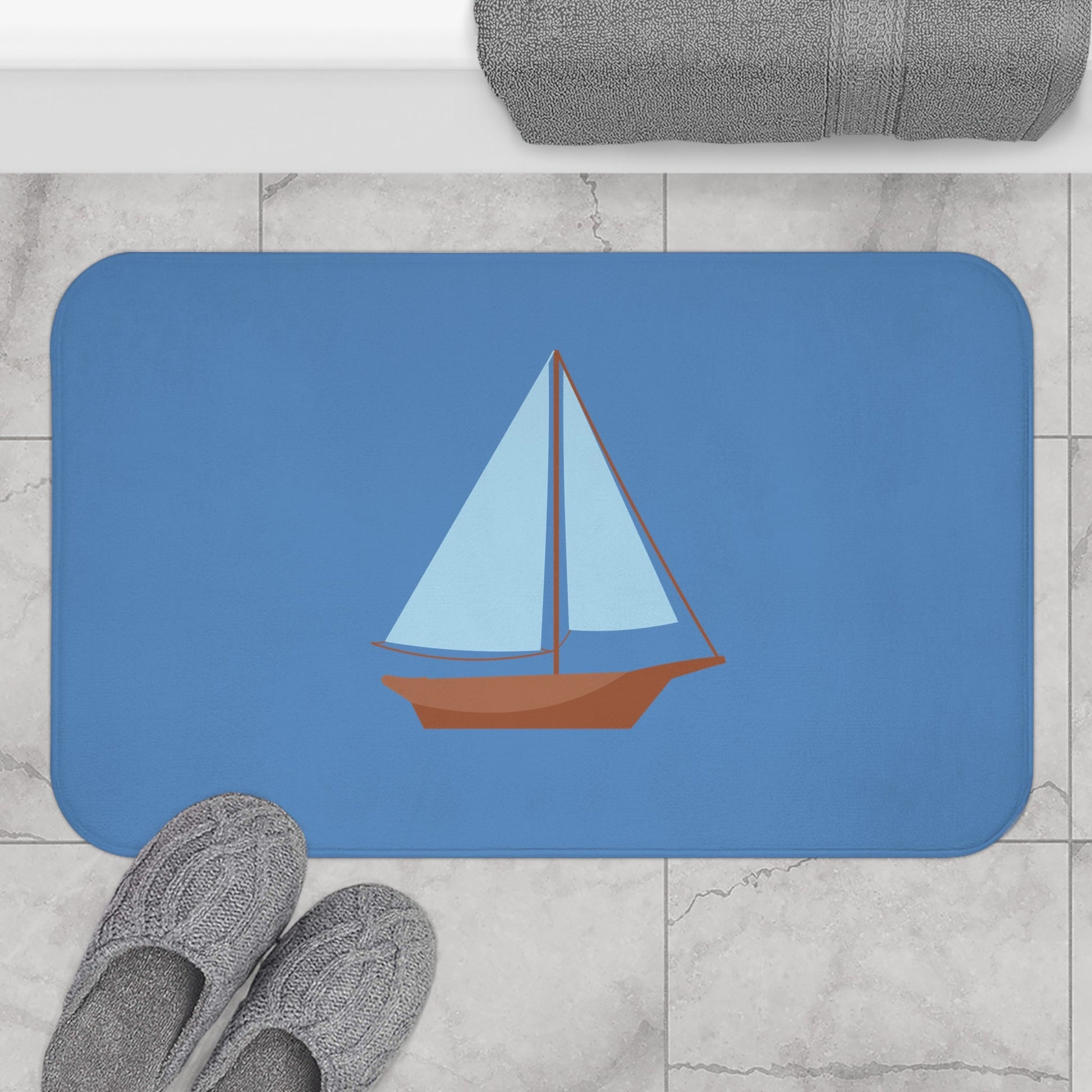 A rectangular blue bath mat with rounded corners featuring a stylized sailboat illustration, shown on a light marble floor with gray slippers and a folded gray towel nearby.
