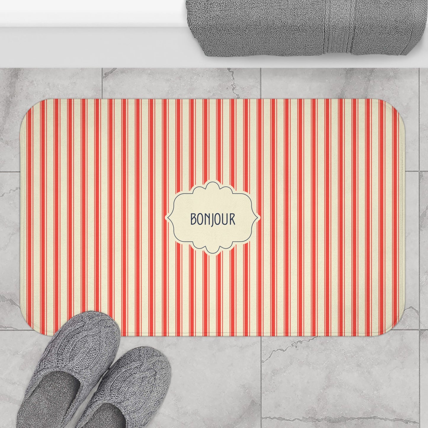 A rectangular bath mat with rounded corners, featuring vertical red and cream stripes and a central cream label that reads 'BONJOUR'. The mat is styled in a bathroom setting on a light gray marble tile floor, with gray slippers at the bottom left and a folded gray towel at the top.
