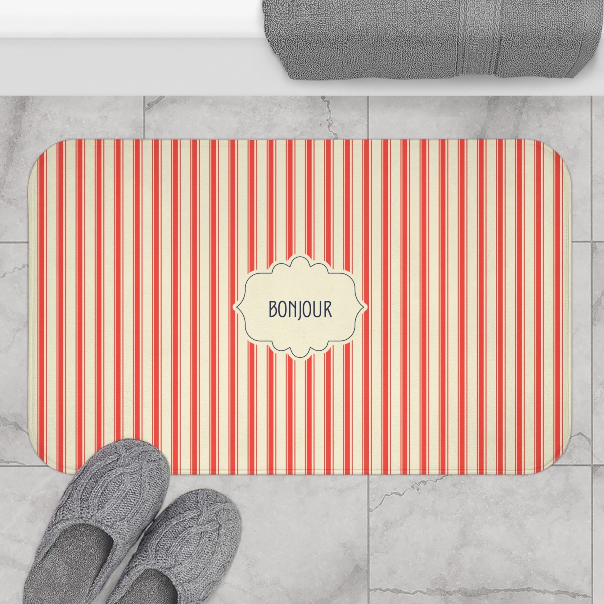 A rectangular bath mat with rounded corners, featuring vertical red and cream stripes and a central cream label that reads 'BONJOUR'. The mat is styled in a bathroom setting on a light gray marble tile floor, with gray slippers at the bottom left and a folded gray towel at the top.