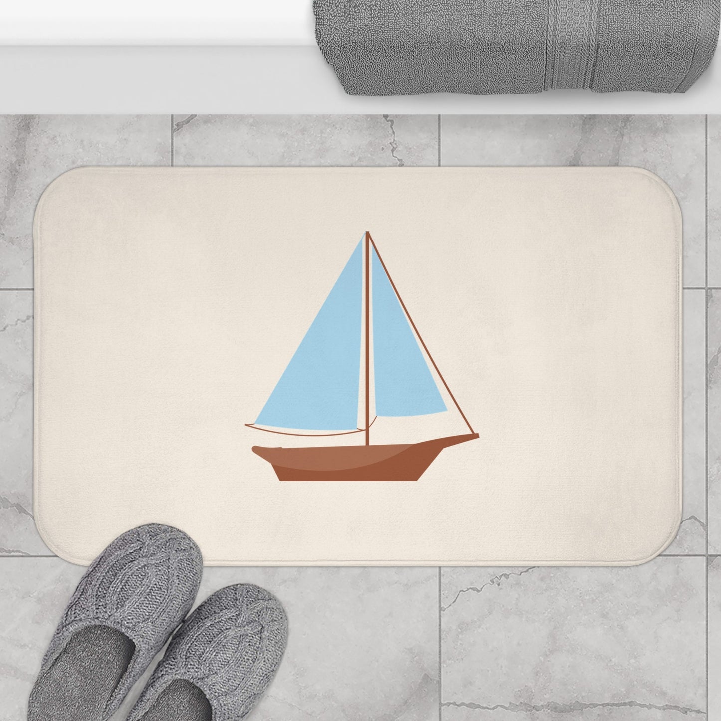 A rectangular bath mat with rounded corners featuring a stylized sailboat illustration, shown on a light gray marble floor with gray slippers and a folded gray towel nearby.