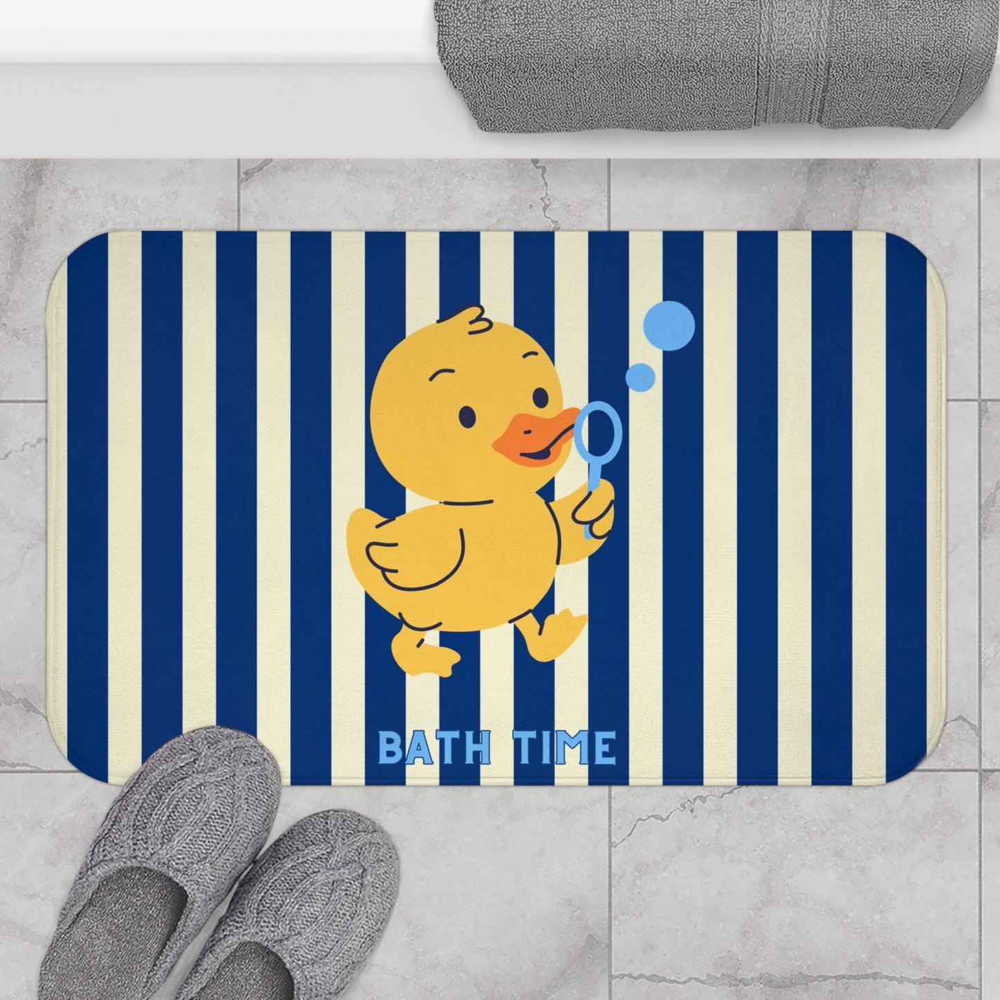 A rectangular bath mat with rounded corners featuring navy and cream vertical stripes, a yellow cartoon duck holding a blue bubble wand, and the words 'BATH TIME' in blue. The mat is shown on a light gray marble floor with gray slippers and a folded gray towel nearby.