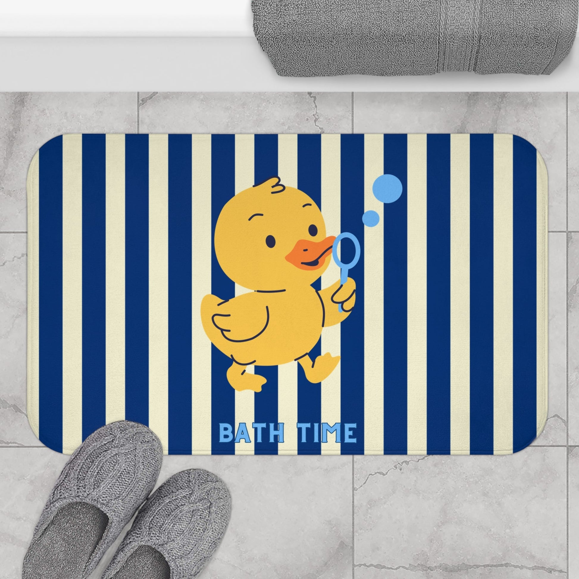A rectangular bath mat with rounded corners featuring navy and cream vertical stripes, a yellow cartoon duck holding a blue bubble wand, and the words 'BATH TIME' in blue. The mat is shown on a light gray marble floor with gray slippers and a folded gray towel nearby.