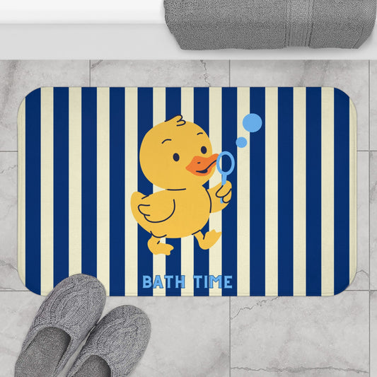 A rectangular bath mat with rounded corners featuring navy and cream vertical stripes, a yellow cartoon duck holding a blue bubble wand, and the words 'BATH TIME' in blue. The mat is shown on a light gray marble floor with gray slippers and a folded gray towel nearby.