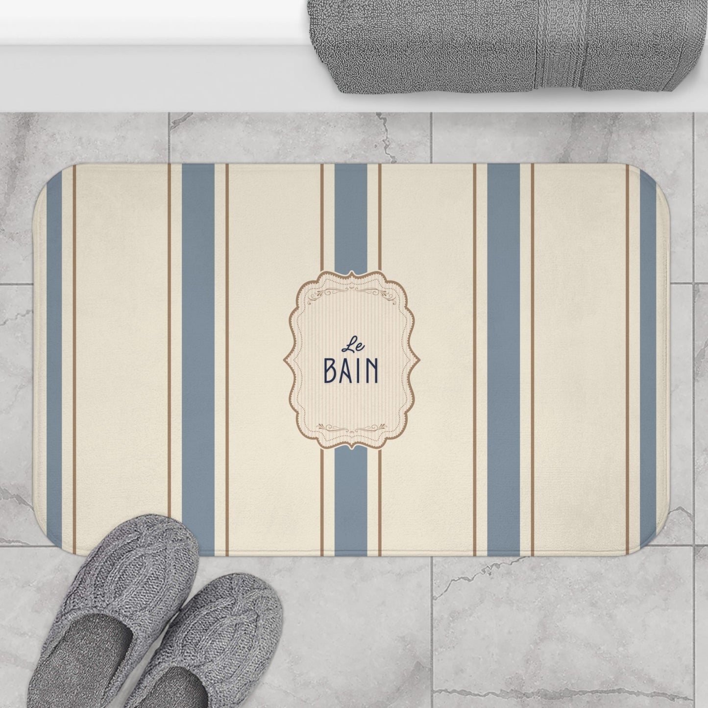 Striped bath mat with 'Le Bain' text on a marble floor with slippers and towel.