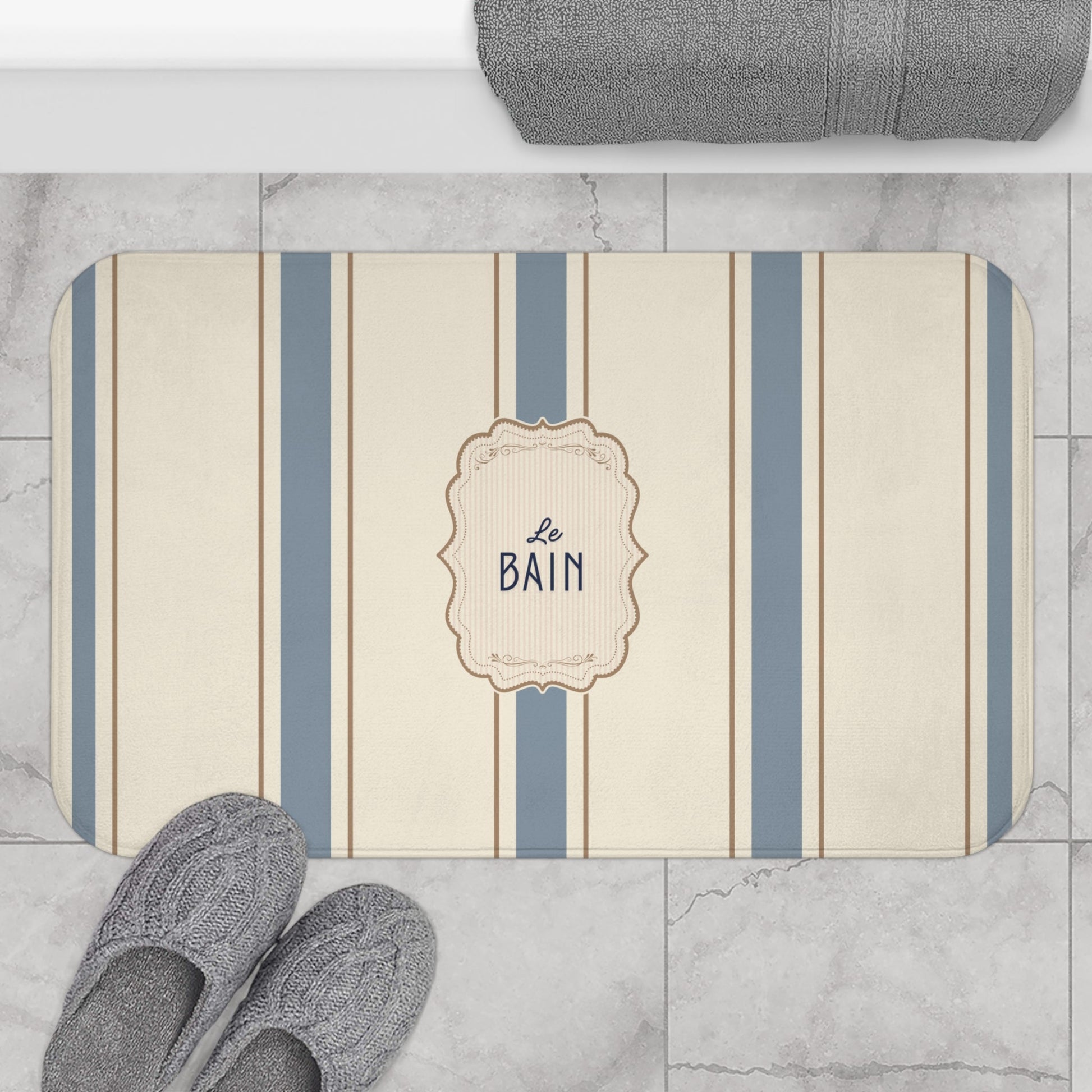 Striped bath mat with 'Le Bain' text on a marble floor with slippers and towel.
