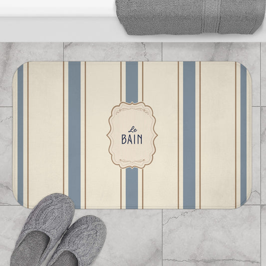 Striped bath mat with 'Le Bain' text on a marble floor with slippers and towel.