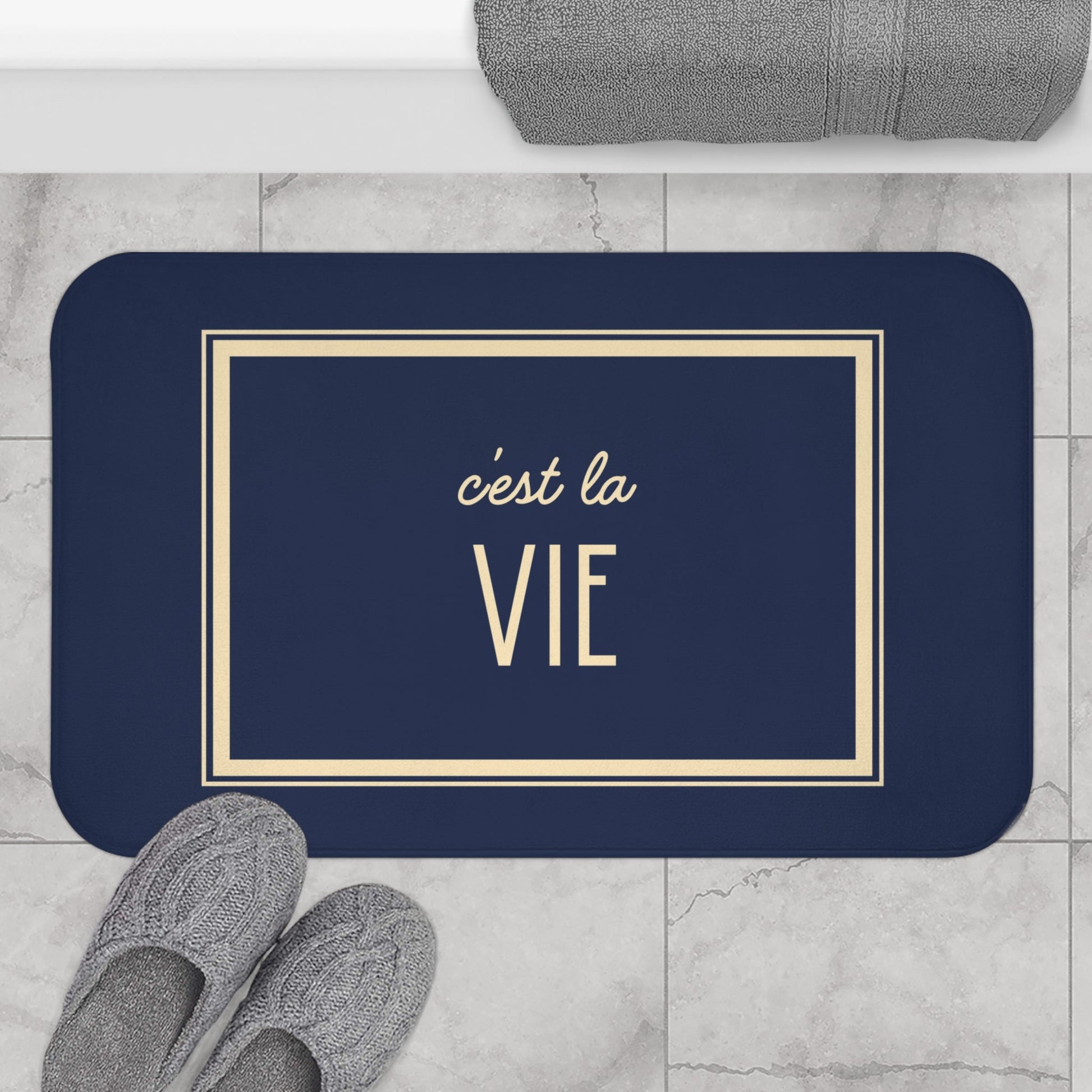 Navy blue mat with 'c'est la Vie' text on a marble floor with slippers and a towel.