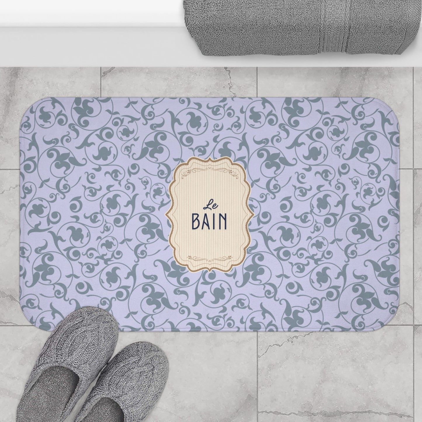 A rectangular bathroom mat with rounded corners, featuring a light lavender background with a dark gray scrollwork pattern and a central cream label that reads 'Le BAIN'. The mat is styled in a bathroom setting on a light gray marble floor, with gray towels and slippers nearby.