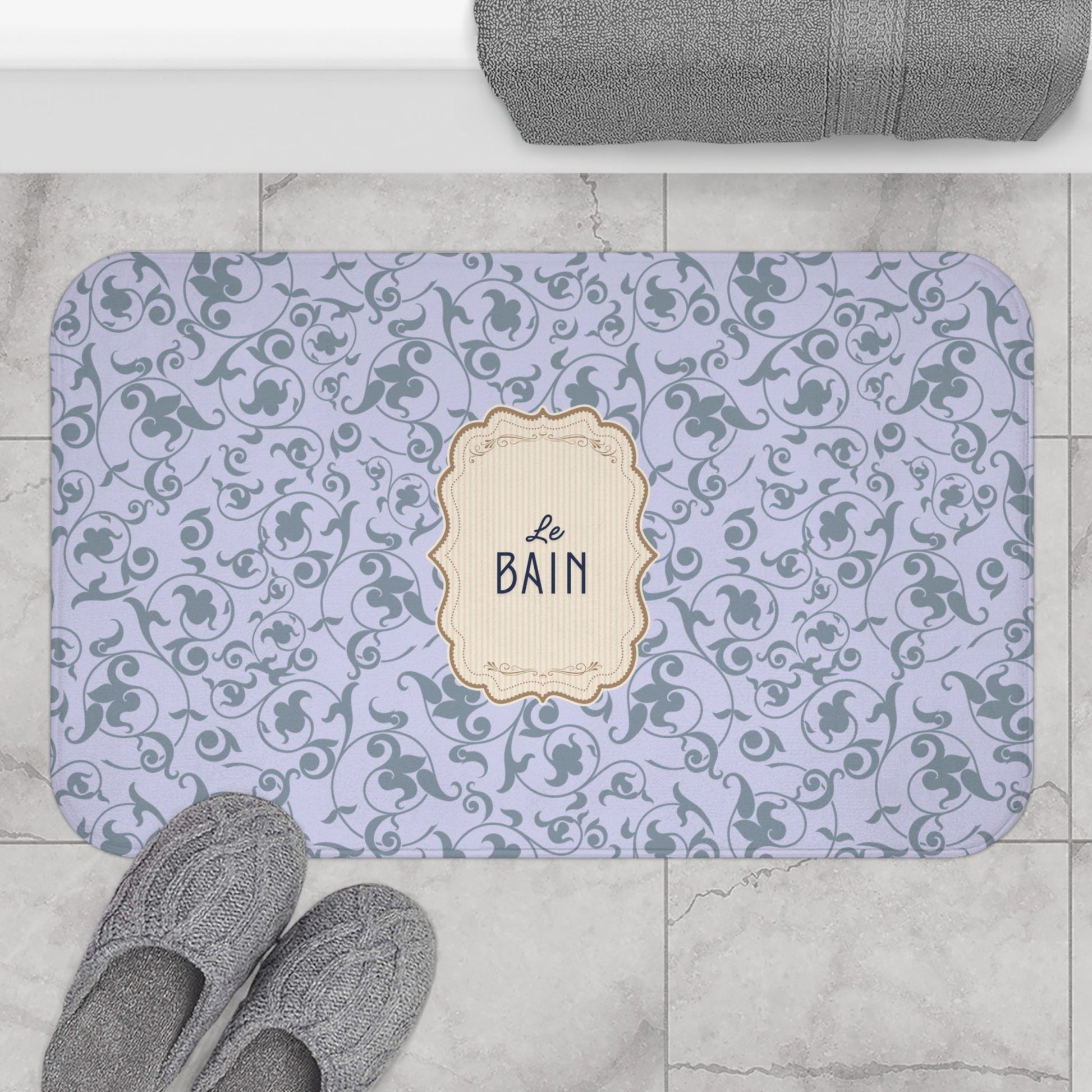 A rectangular bathroom mat with rounded corners, featuring a light lavender background with a dark gray scrollwork pattern and a central cream label that reads 'Le BAIN'. The mat is styled in a bathroom setting on a light gray marble floor, with gray towels and slippers nearby.