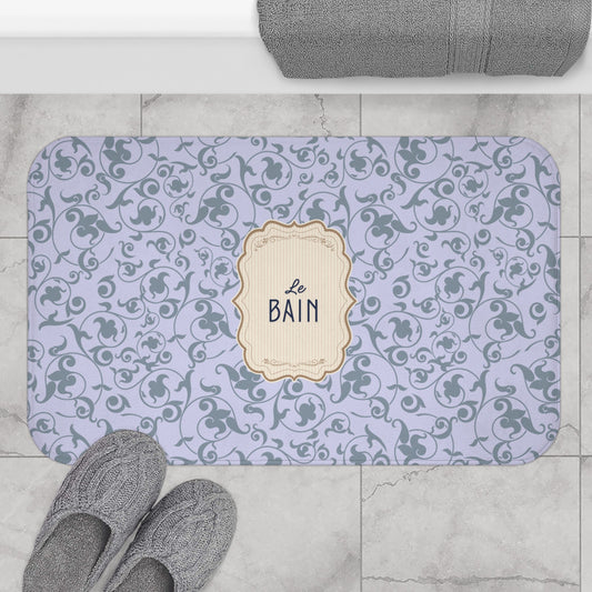 A rectangular bathroom mat with rounded corners, featuring a light lavender background with a dark gray scrollwork pattern and a central cream label that reads 'Le BAIN'. The mat is styled in a bathroom setting on a light gray marble floor, with gray towels and slippers nearby.