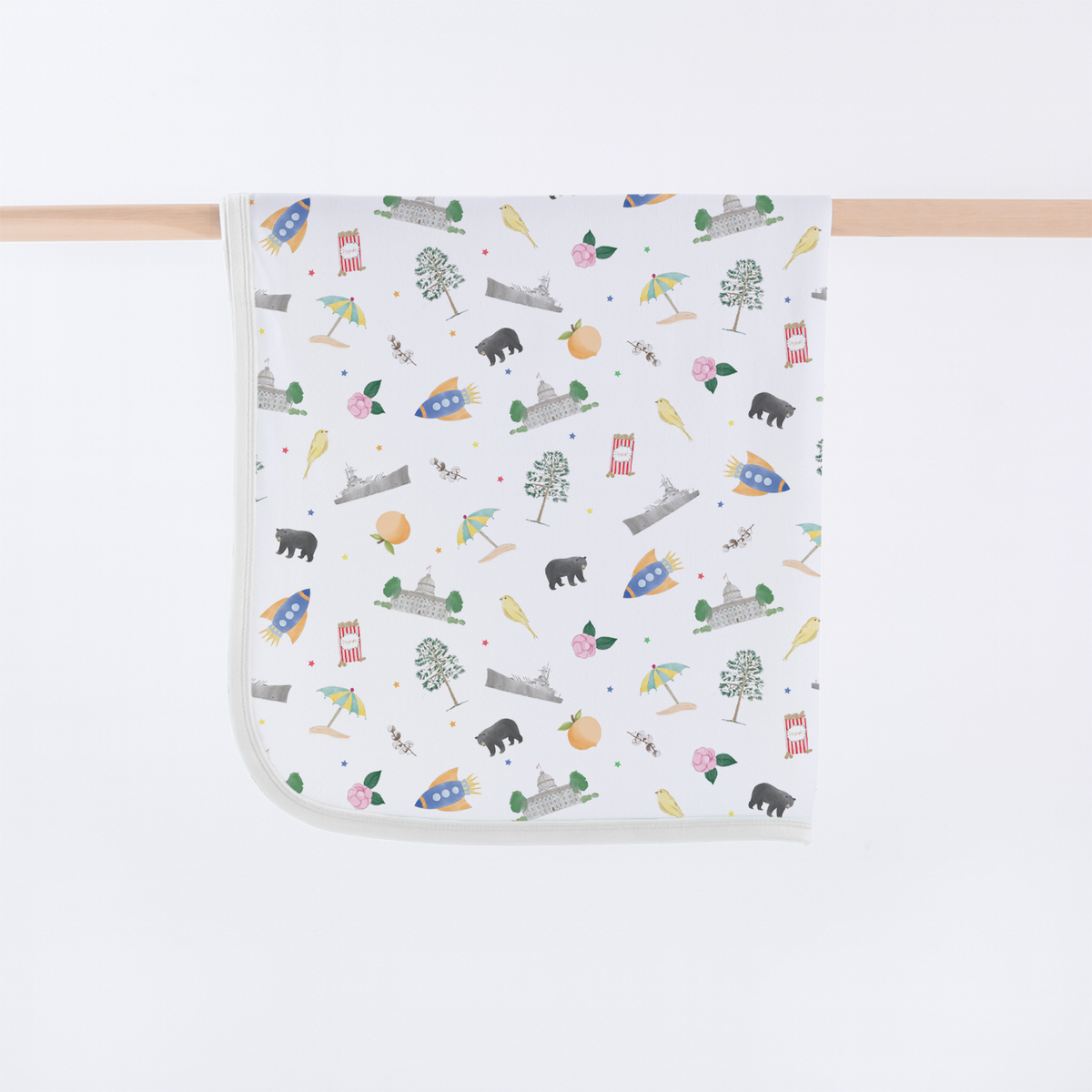 A square white baby blanket with rounded corners, featuring a colorful cartoon pattern of rockets, trees, owls, bears, and houses, hanging from a light wood dowel against a white background.