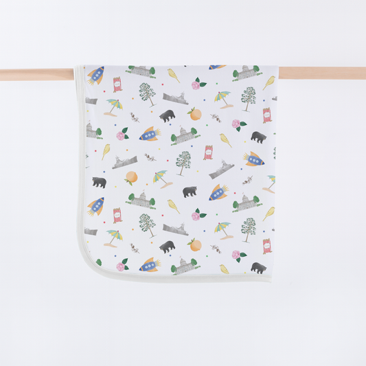 A square white baby blanket with rounded corners, featuring a colorful cartoon pattern of rockets, trees, owls, bears, and houses, hanging from a light wood dowel against a white background.