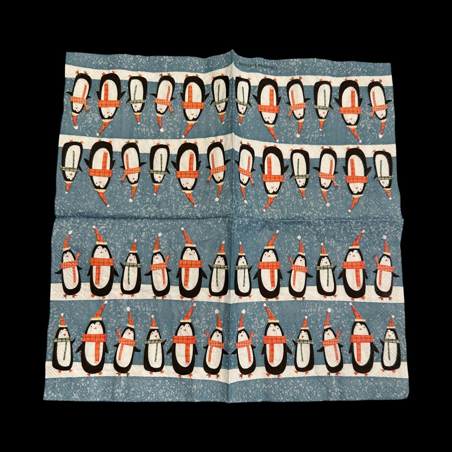 Open unfolded Cocktail size decoupage napkin featuring rows of cute penguins wearing scarves and Santa hats on a snowy blue background for holiday crafts and decor.