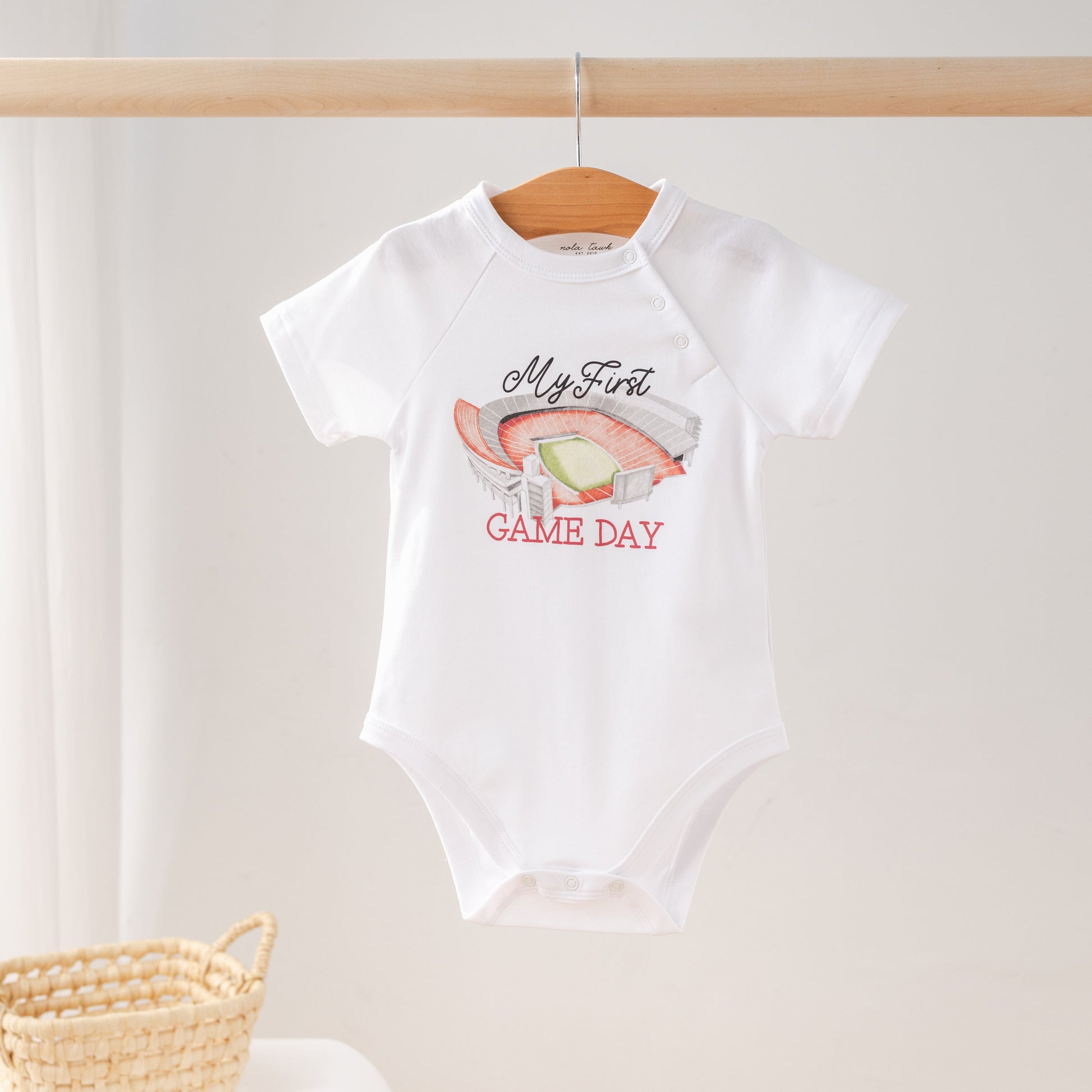 White baby onesie with 'My First Game Day' text and graphics on a wooden hanger.