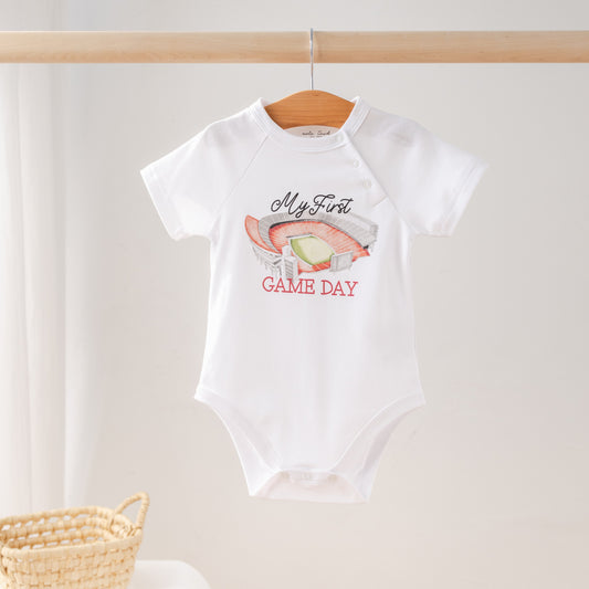 White baby onesie with 'My First Game Day' text and graphics on a wooden hanger.