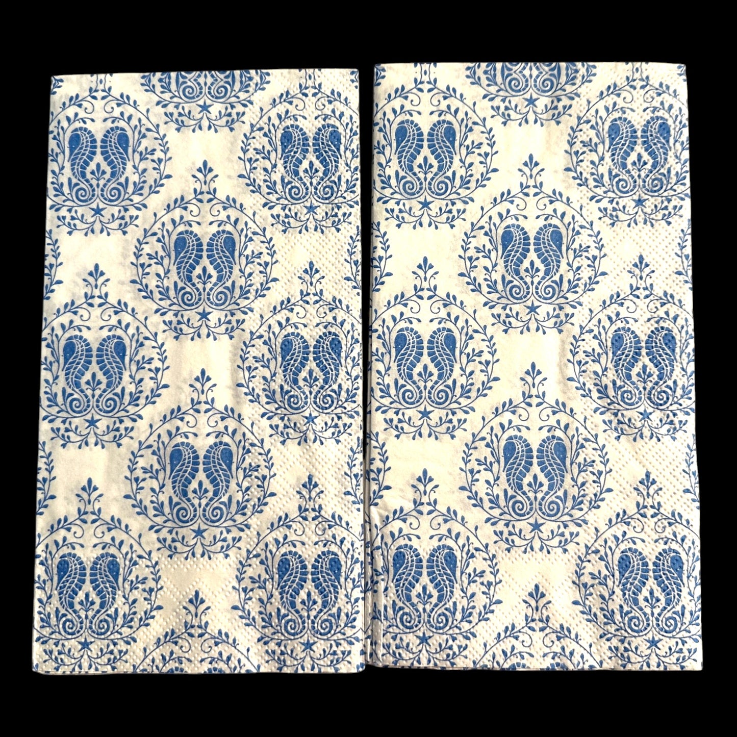 Two guest-size decoupage paper napkins featuring a detailed all-over print of classic paisley teardrops with intricate interior scrollwork and trailing vine and leaf sprigs in rich cobalt and periwinkle blue on a soft warm white background, photographed on a light grey surface.