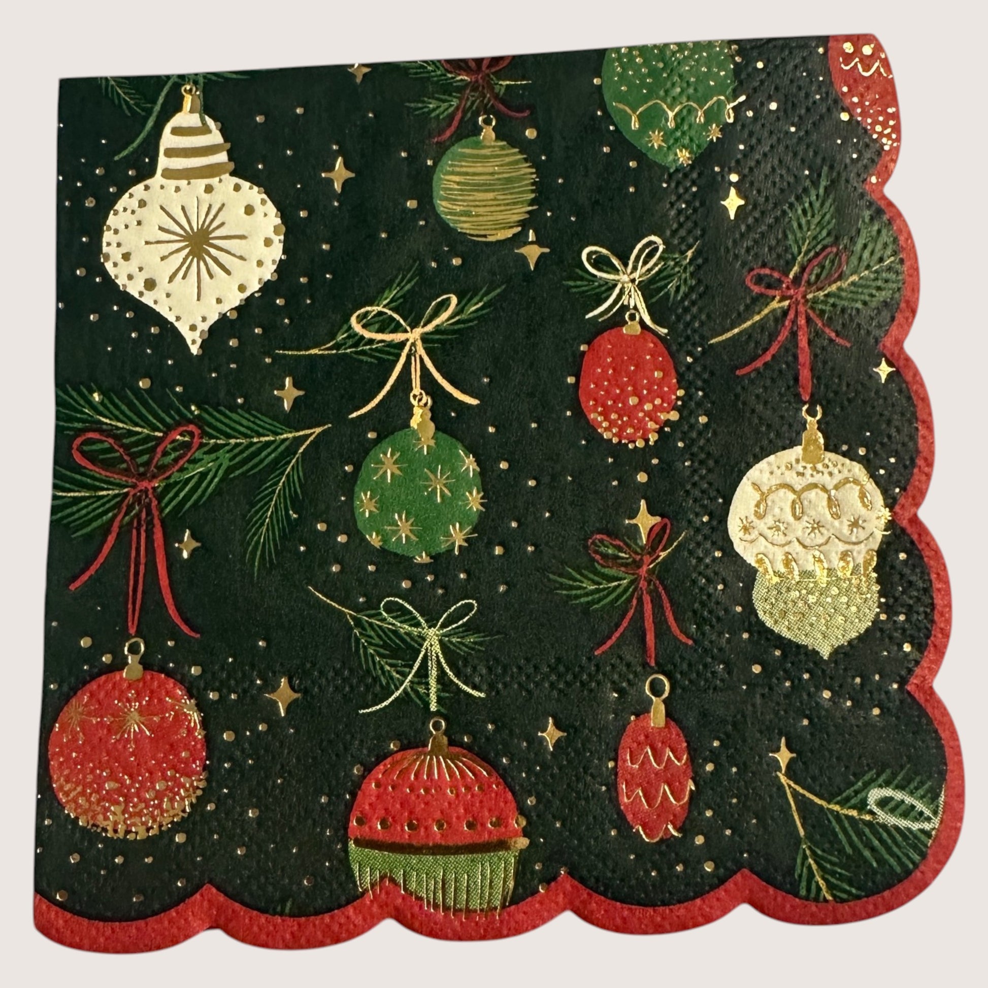 Cocktail-size decoupage paper napkin featuring an all-over print of hanging Christmas ornaments in deep red forest green cream and gold with gold foil detailing suspended from pine sprigs with ribbon bows on a deep forest green background with gold star accents and a red scalloped edge border, photographed on a light grey surface.