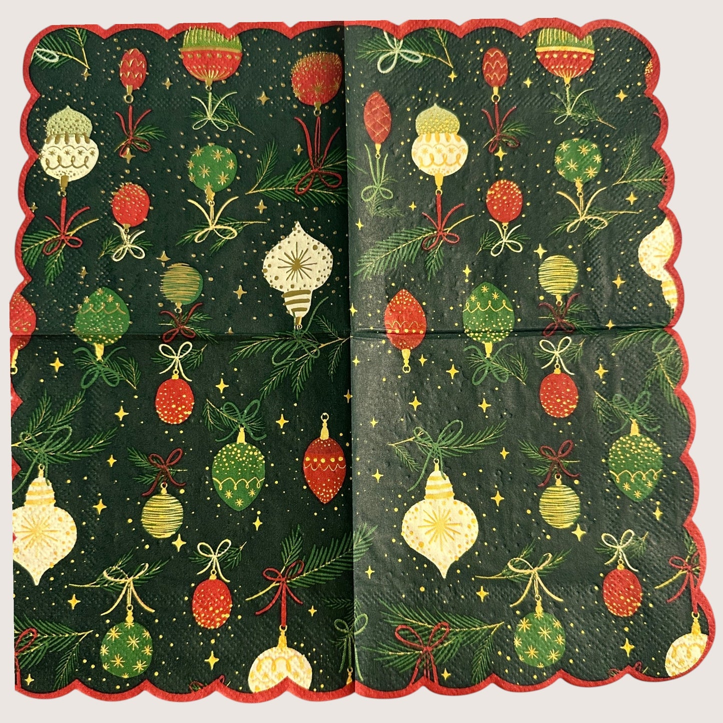Cocktail-size decoupage paper napkin featuring an all-over print of hanging Christmas ornaments in deep red forest green cream and gold with gold foil detailing suspended from pine sprigs with ribbon bows on a deep forest green background with gold star accents and a red scalloped edge border, photographed on a light grey surface.