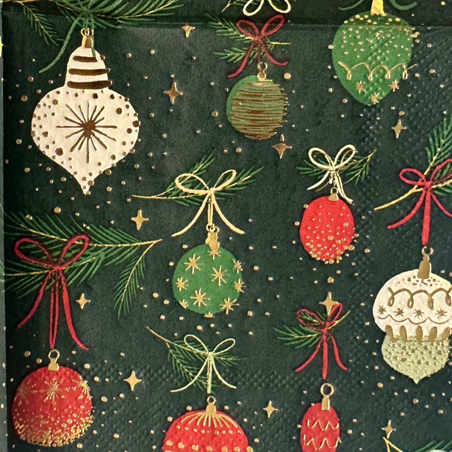 Cocktail-size decoupage paper napkin featuring an all-over print of hanging Christmas ornaments in deep red forest green cream and gold with gold foil detailing suspended from pine sprigs with ribbon bows on a deep forest green background with gold star accents and a red scalloped edge border, photographed on a light grey surface.