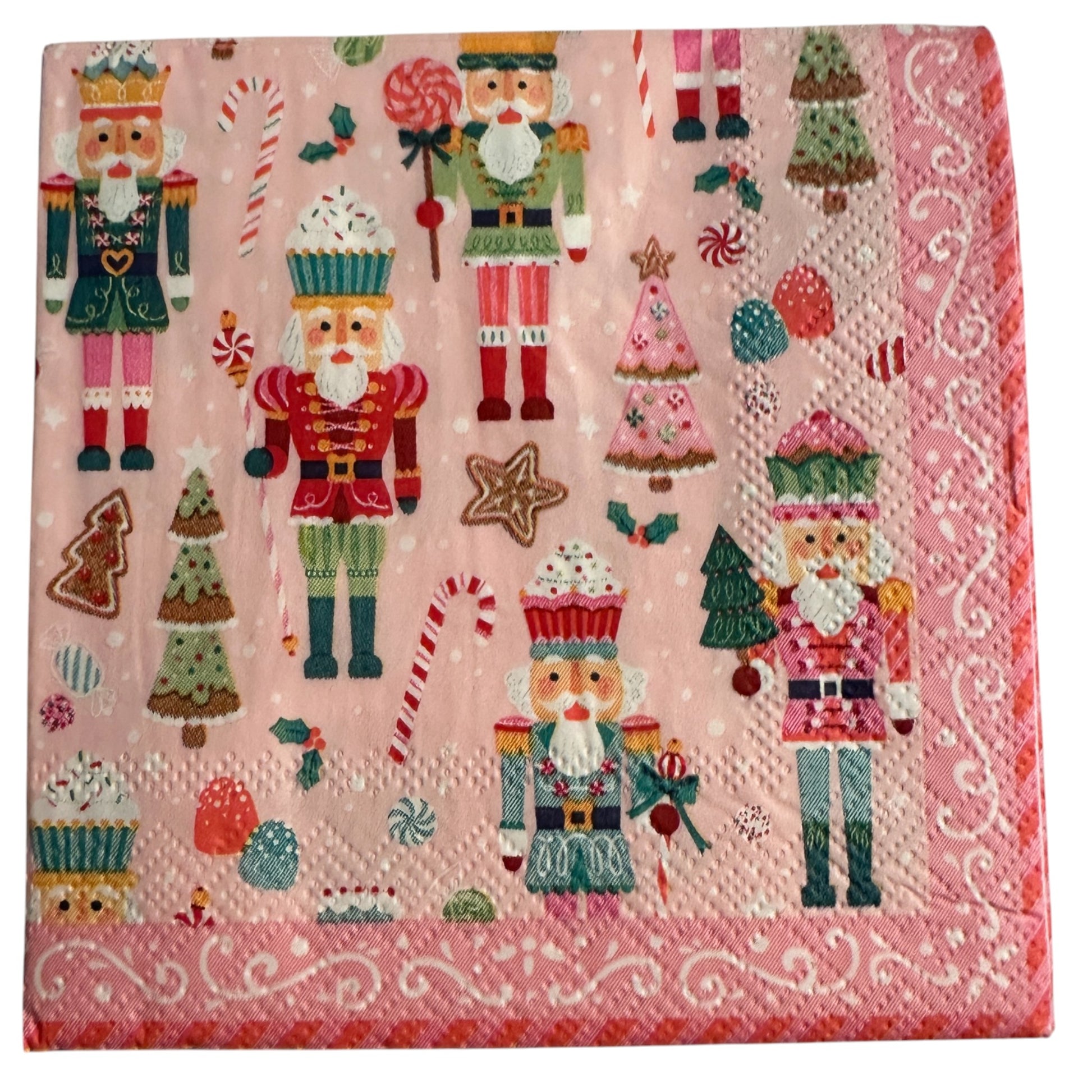 Cocktail-size decoupage paper napkin featuring a lively all-over print of colorful nutcracker soldiers in teal red green and pink uniforms with candy canes lollipops cupcake nutcrackers gingerbread trees pink Christmas trees and star cookies on a soft bubblegum pink background with a white scrollwork lace border, photographed on a light grey surface.