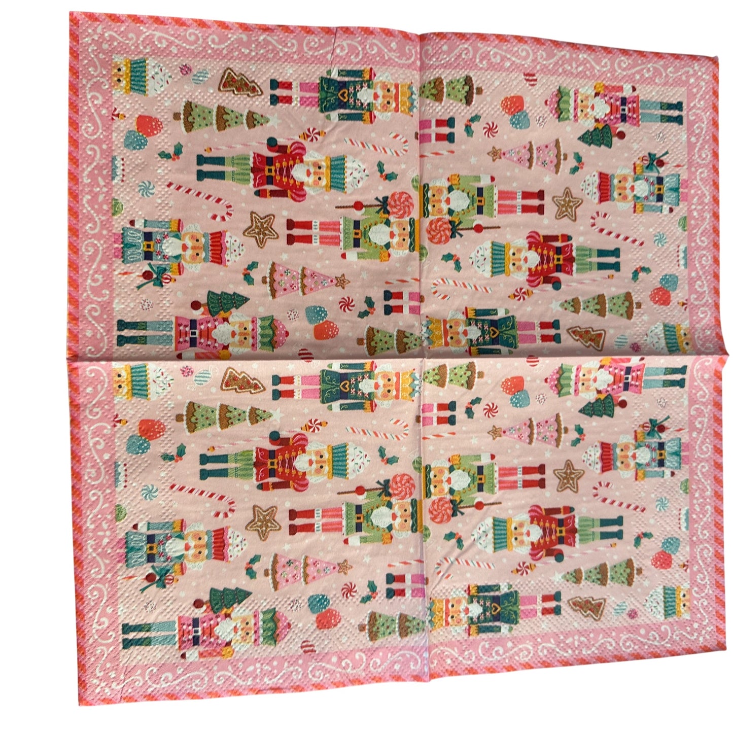 Cocktail-size decoupage paper napkin featuring a lively all-over print of colorful nutcracker soldiers in teal red green and pink uniforms with candy canes lollipops cupcake nutcrackers gingerbread trees pink Christmas trees and star cookies on a soft bubblegum pink background with a white scrollwork lace border, photographed on a light grey surface.