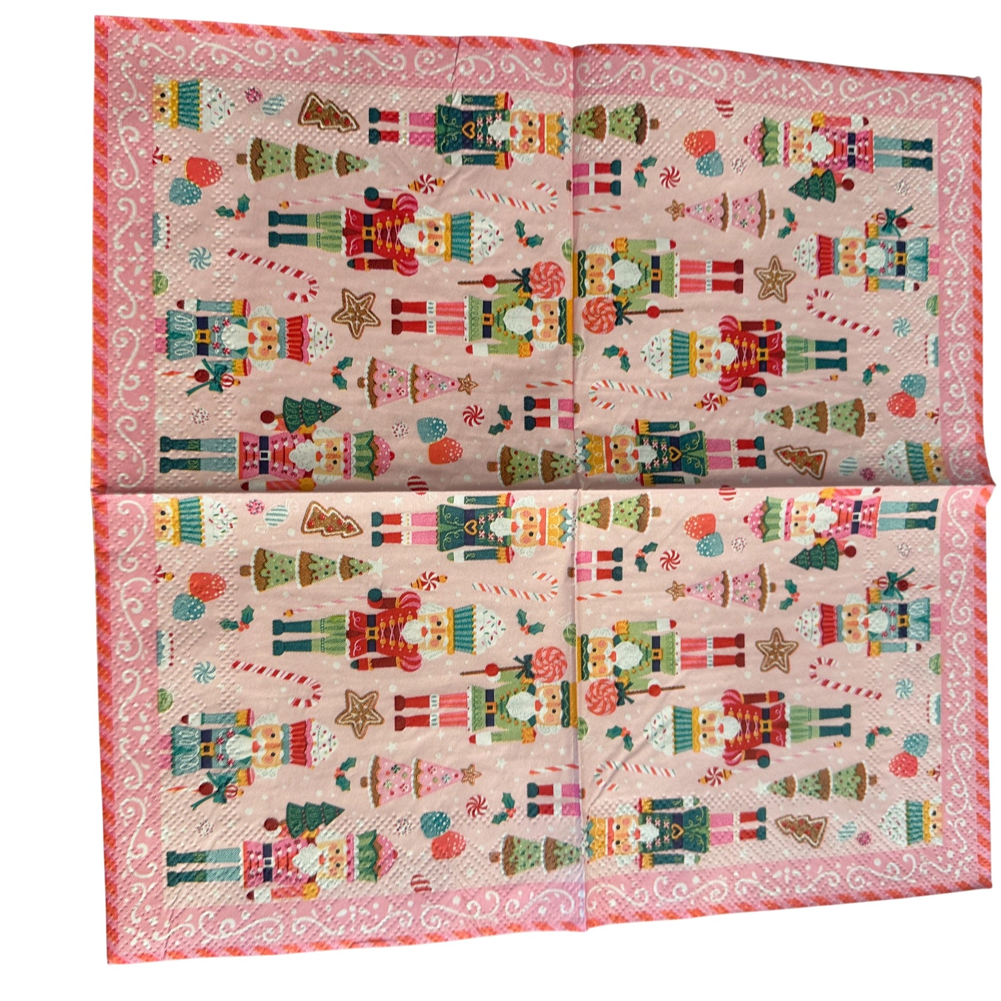 Cocktail-size decoupage paper napkin featuring a lively all-over print of colorful nutcracker soldiers in teal red green and pink uniforms with candy canes lollipops cupcake nutcrackers gingerbread trees pink Christmas trees and star cookies on a soft bubblegum pink background with a white scrollwork lace border, photographed on a light grey surface.