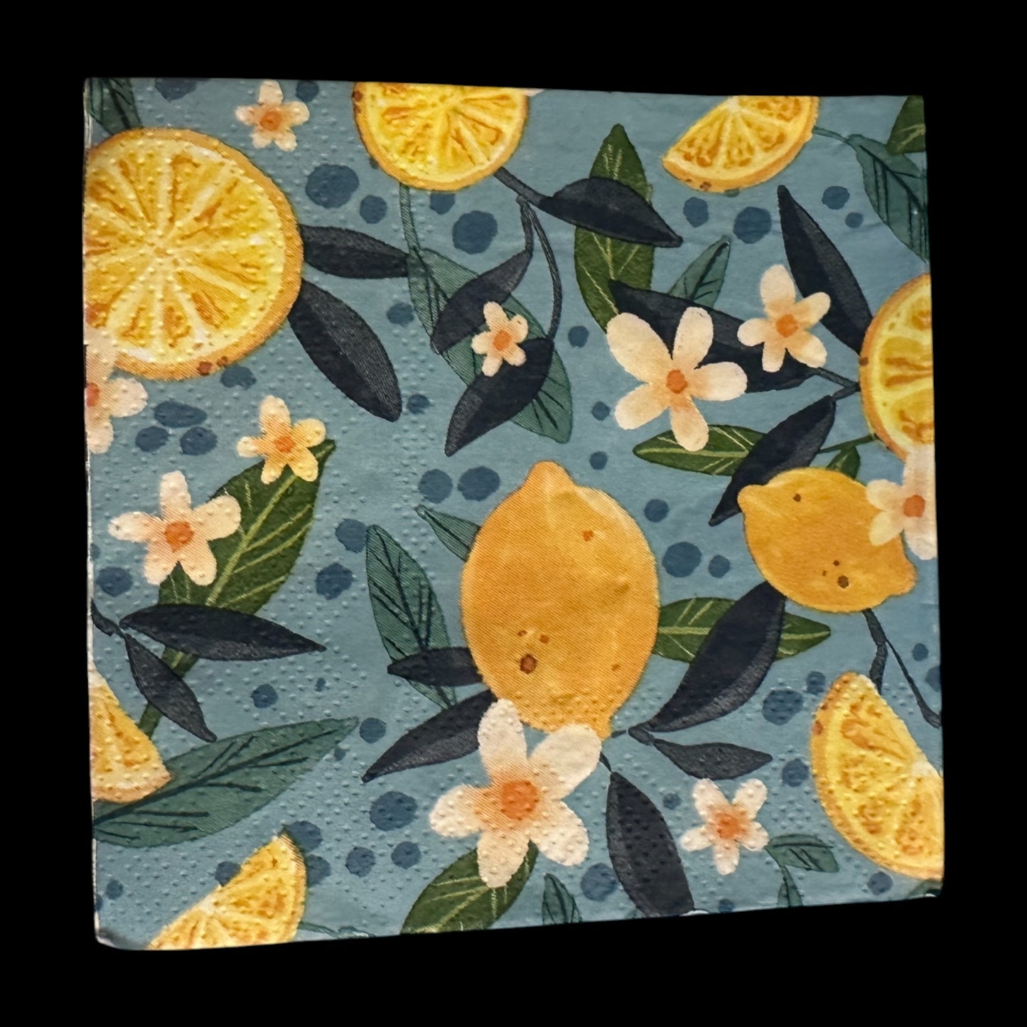 Cocktail-size decoupage paper napkin featuring a lush all-over botanical print of golden whole lemons and halved citrus slices with white orange blossom flowers and rich dark and forest green leaves on a soft dusty blue background with subtle dot accents, photographed on a black surface.