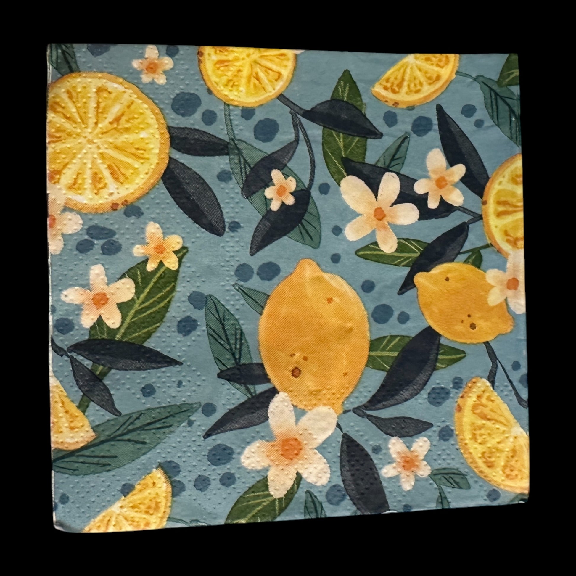 Cocktail-size decoupage paper napkin featuring a lush all-over botanical print of golden whole lemons and halved citrus slices with white orange blossom flowers and rich dark and forest green leaves on a soft dusty blue background with subtle dot accents, photographed on a black surface.